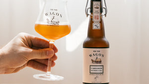 A hand holding a glass filled with kombucha, alongside a bottle of On The Wagon Cardamom & White Tea kombucha