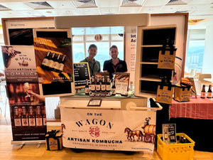 A kombucha booth display at an indoor event, featuring branded signs, bottles, a counter with two people behind it, and a large banner reading 'On the Wagon Artisan Kombucha' with a horse-drawn wagon illustration.