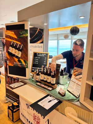 A man in a navy blue shirt works behind a display counter filled with kombucha bottles, signs, and a small sign reading 'Artisanal Kombucha'.