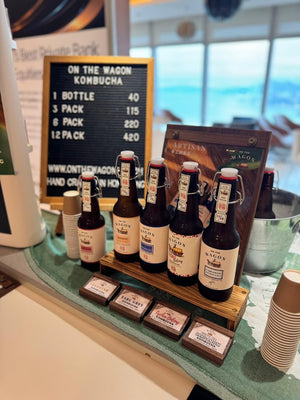 A display of On the Wagon kombucha bottles and boxes on a counter, with a black letterboard sign showing pricing and a window in the background.