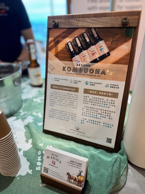 A wooden-framed sign displaying a kombucha product photo and descriptive text, standing on a green cloth-covered table with a small business card in front. A person and bar counter are blurred in the background.
